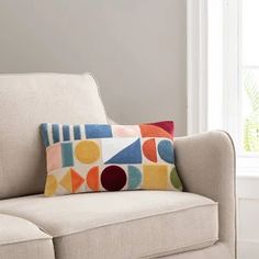 a colorful pillow sitting on top of a beige couch next to a white chair and window