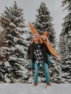 two people standing in the snow with their arms around each other and trees behind them