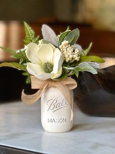 Pretty winter mason jar floral arrangement is the perfect holiday accent. A single beautiful  magnolia flower sits nestled among a bouquet of snow kissed lambs ear, italian ruscus and fern. Elegant with just a hint of winter sparkle, you'll want to display this beauty all season long. Need a holiday gift? This piece is perfect! Pint sized mason jar has been hand painted a pretty winter white, using environmentally friendly and toxin free mineral paint.  Finished on all sides, but meant to be viewed from the front, this winter arrangement measures approximately 10 1/2" high by 11" wide, the perfect size for so many spaces! You can be confident in the quality of florals you will receive, as all of the materials used in my floral arrangements have been hand selected by myself for their qualit Winter Greenery Centerpiece, January Table Centerpieces, Winter Floral Arrangements Home, January Flower Arrangements, Winter Centerpieces For Table, Magnolia Centerpiece, January Flower, Mason Jar Arrangements, Winter Flower Arrangements