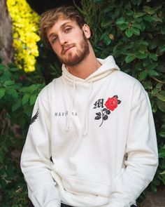 a man with a beard wearing a white hoodie standing in front of some bushes