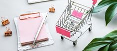 a shopping cart next to a pink notepad and pen on a white table with green leaves