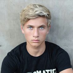 a young man with blonde hair and blue eyes wearing a black shirt looking at the camera