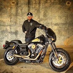 a man standing next to a black motorcycle in a parking lot with a wall behind him