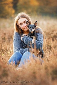 a woman holding a small dog in her arms while sitting on the ground with tall grass