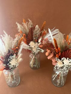 three vases filled with flowers and feathers on a brown tableclothed surface in front of a wall