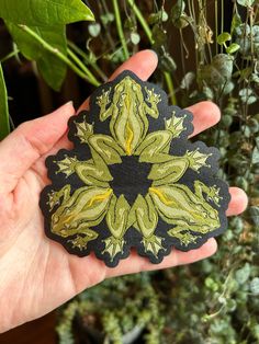 a hand holding up a green and black patch with leaves on it in front of some plants