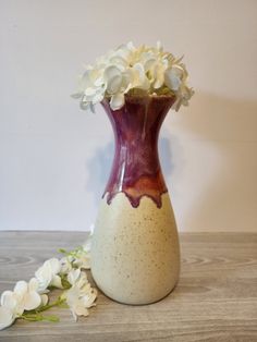 a white and purple vase with flowers in it on a wooden table next to a wall