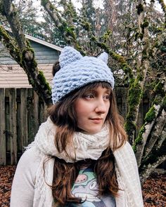 a woman wearing a knitted hat and scarf in front of a tree with leaves on the ground