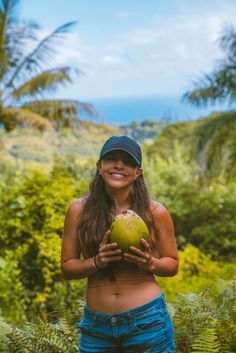 a young woman holding a coconut in her hands and smiling at the camera, with tropical vegetation behind her