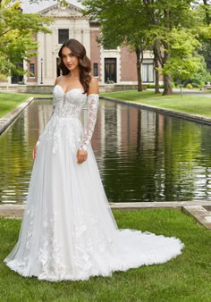 a woman in a white wedding dress standing on the grass near a body of water