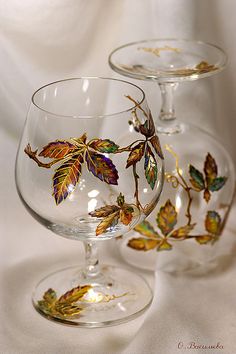 two wine glasses with leaves painted on the glass are sitting on a white tablecloth