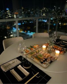 a table topped with sushi and candles next to a glass of wine in front of a window