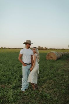 a man and woman standing next to each other in a field with hay bales