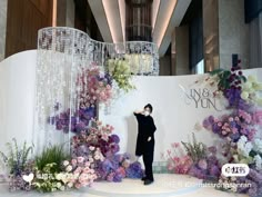 a man standing on top of a stage surrounded by purple and white floral decorations in front of a chandelier