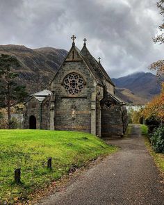 an old church in the middle of a grassy field with mountains in the background and cloudy skies