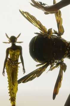 a woman is walking down the runway with gold wings on her head and body, while two hands are reaching out towards it