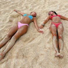 two women in bikinis laying on the sand with their arms around each other,