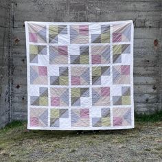 a quilt hanging on the side of a building with grass in front of it and a brick wall behind it