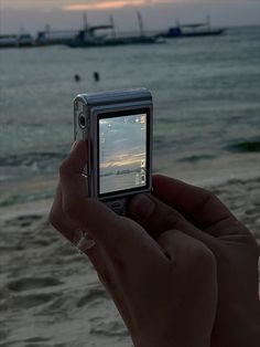 a person holding up a cell phone to take a photo on the beach with boats in the background