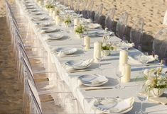 a long table set up with white plates and silverware on the beach for a wedding reception