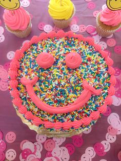 a birthday cake with sprinkles and smiley face surrounded by cupcakes