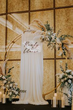 a wedding arch decorated with white flowers and greenery is surrounded by candles in front of a gold wall