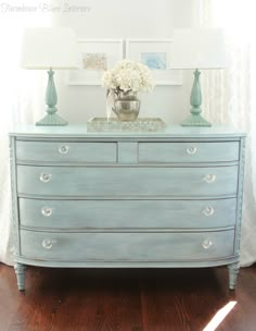 a blue dresser with white flowers on top and two lamps on the side, in front of a window