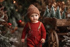 a small child in a red jumpsuit and knitted hat standing next to christmas trees