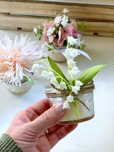 a hand holding a jar filled with flowers on top of a white table next to other vases