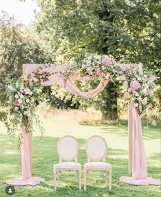 two white chairs sitting under an arch with pink flowers and greenery on the side