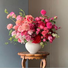 a white vase filled with pink and red flowers on top of a wooden table next to a gray wall