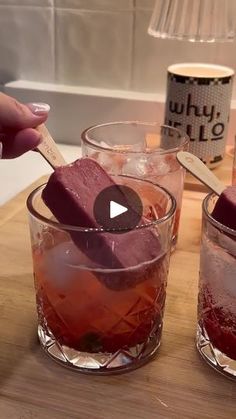 two glasses filled with liquid and ice cubes on top of a wooden table next to a lamp