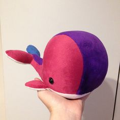 a hand holding a pink and purple whale stuffed animal in front of a white wall