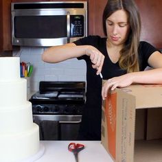 a woman cutting a cake with scissors on the counter next to a box and knife