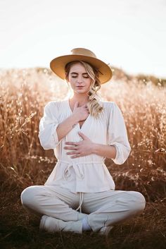 a woman wearing a hat sitting in the middle of a field with her hands on her chest