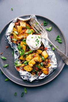 a plate topped with foil covered in potatoes and bacon next to a knife and fork