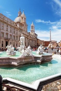 a fountain in front of a large building with statues on it's sides and people walking around