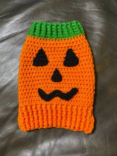 an orange knitted pumpkin hat with black eyes and a green top on a leather surface