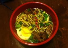 a red bowl filled with noodles, meat and veggies on top of a wooden table