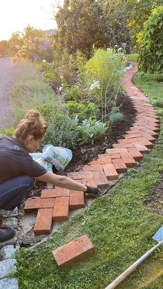 a woman laying bricks on the ground in front of a garden area with grass and flowers