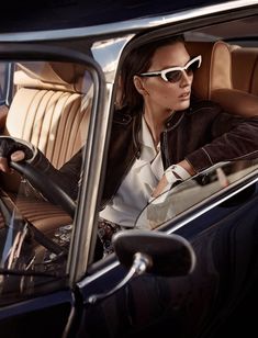 a woman sitting in the driver's seat of a car wearing sunglasses and a leather jacket