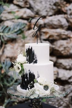 a white and black wedding cake on a glass stand with succulents, greenery and rocks in the background