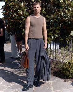 a man standing in front of a tree holding a brown bag and wearing black shoes