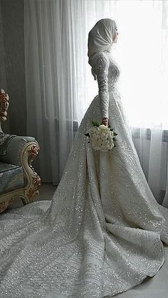 a woman in a wedding dress standing next to a chair and window with curtains behind her