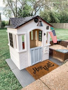 a toy house with an american flag on the front door and side porch, sitting in a yard