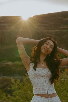 a woman standing in the grass with her arms behind her head and hands on her hair