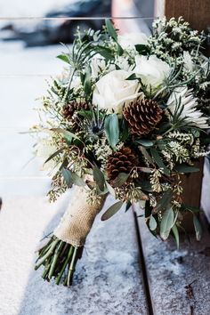 a bridal bouquet with white flowers and pine cones is sitting on a wooden bench