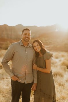 a man and woman standing together in the desert with mountains in the background at sunset