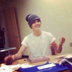 a young man sitting at a table in front of a laptop computer and wearing a beanie