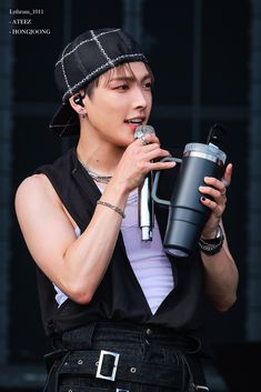 a male in a black vest and hat is holding a coffee cup while singing into a microphone